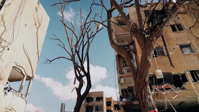 An upward facing wide shot, at a heavy missile falling site, during the Israel Iran war, where the buildings are broken, trees are burnt and few birds are flying across. Arad, Israel