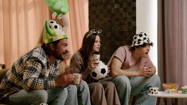 Group of friends wearing soccer hats and holding cups cheer enthusiastically while watching a soccer match on television in a cozy living room setting
