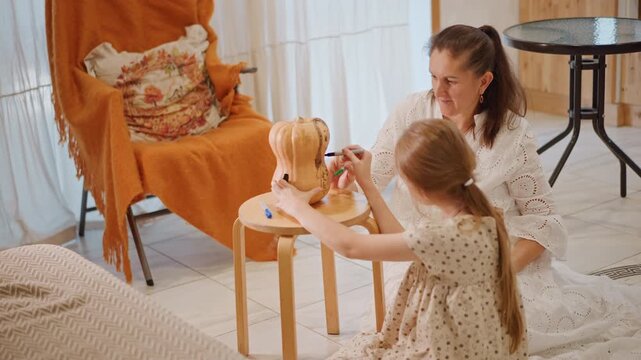 White mother and daughter decorate pumpkin at small round table, playful craft session with paint and tiny tools, cozy armchair draped in orange throw, mom as craft teacher encouraging imaginative