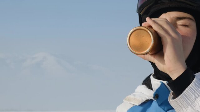 Young man in ski helmet and goggles on helmet drinking hot tasty beverage from orange thermos in mountains. Young man happy to be in mountains and ride snowboard. Copy space. Active lifestyle.