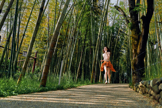 Woman running through a bamboo forest on a shaded path and nature trail, smile visible as she jogs in casual dress amid green woodland scenery, outdoor exercise and freedom.