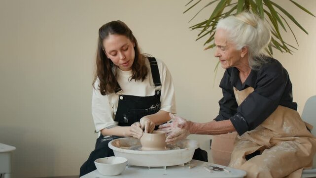 Elderly and young women working together at potters wheel, shaping piece and assisting each other in workshop