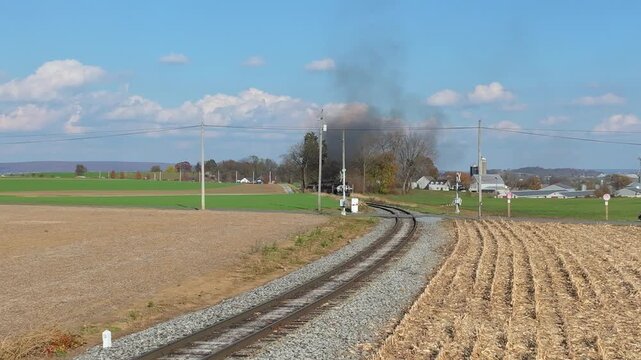 A steam train moves along the tracks through open fields. Smoke rises from the engine as it approaches the camera. The sun shines and the landscape is green and brown.