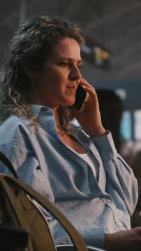 Woman Sits Comfortably In Airport Lounge, Enjoying Coffee While Having Cheerful Phone Conversation. Stress Free Travel Experience, Positive Emotions, Staying Connected With Loved Ones. Vertical Shot.