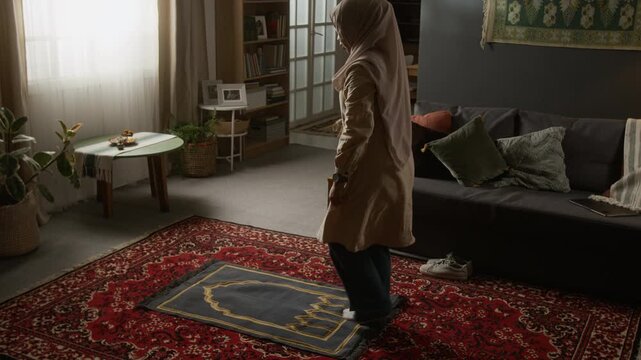 Wide angle shot of young Muslim woman wearing modest clothing staring daily prayer ritual and laying out prayer mat at home with traditional decor elements, copy space