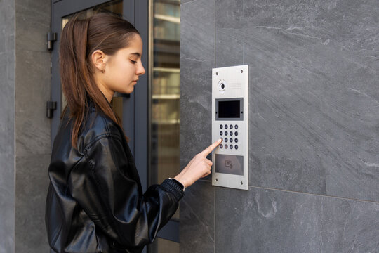 Young woman using intercom at apartment entrance. Young woman presses a video intercom keypad at a modern apartment entrance, illustrating residential security, access control, and urban living.