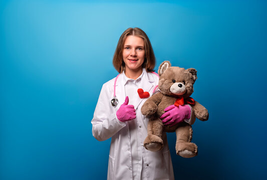 Pediatrician in a white coat holds a teddy bear and gives a thumbs up. Portrait of a female doctor with a plush toy on a blue background