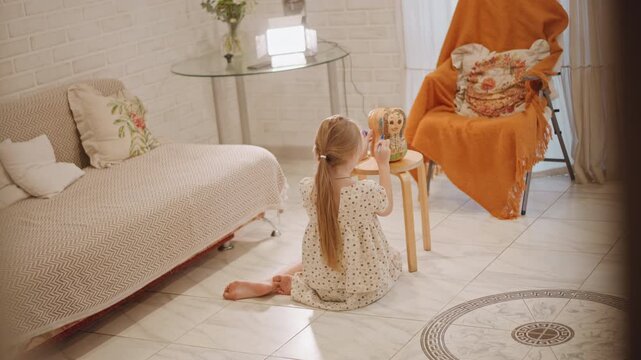 Little white girl playing with doll in cozy sunlit room, kneeling on rug beside couch and orange chair, tenderly arranging toy, vintage floral pillows and knitted blanket, warm morning light, quiet