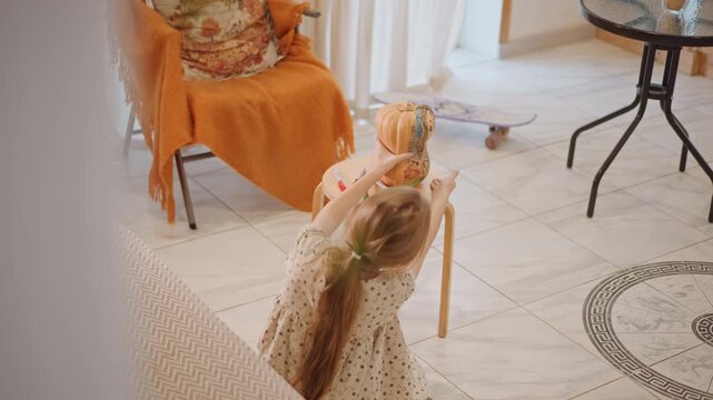 White girl offering pumpkin pie to doll, cozy living room scene with orange throw on armchair, wooden stool holding decorative pie, tiled floor and patterned rug, braided hair and polka dot dress,