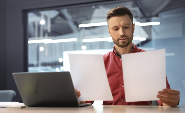 A man sits at a desk with a laptop and holds two documents. He appears focused as he looks closely at the papers. The setting is a modern office with bright lights.