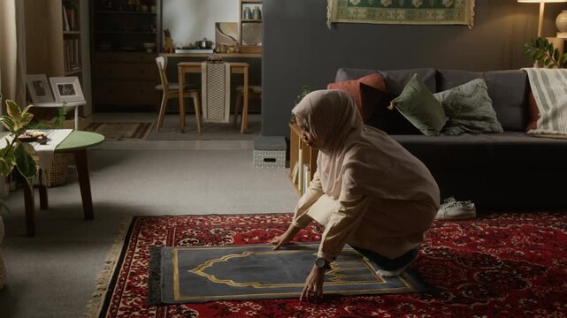 Wide angle shot of young Muslim woman finishing daily prayer and standing up folding prayer mat in modern home interior with traditional decor lit by cinematic lighting, copy space