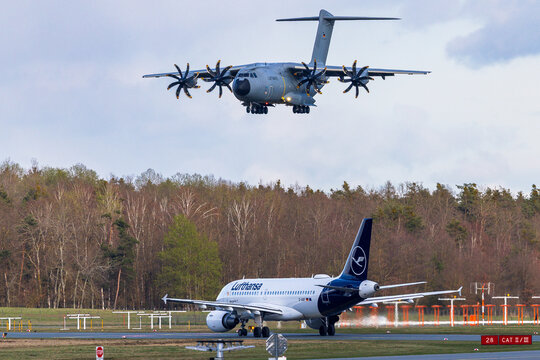 Ein Airbus A319 der Lufthansa im Vordergrund &uuml;ber das Flughafengel&auml;nde, w&auml;hrend im Hintergrund ein Milit&auml;rtransportflugzeug vom Typ Airbus A400M der Luftwaffe im Anflug zu sehen ist. 