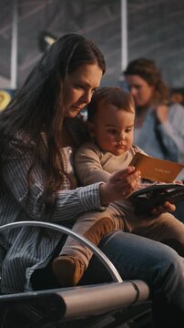 Focused Mother Sits in Airport Terminal, Holding Baby While Children Both Look Smartphone Screen and Boarding Documents. Digital Distractions For Children and Family Travel Lifestyle. Vertical Shot.