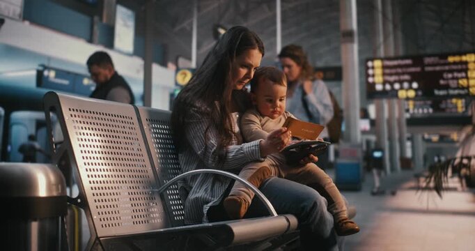 Focused Mother Sits in Airport Terminal, Holding Baby While Children Both Look at Smartphone Screen and Boarding Documents. Concept of Digital Distractions For Children and Family Travel Lifestyle.