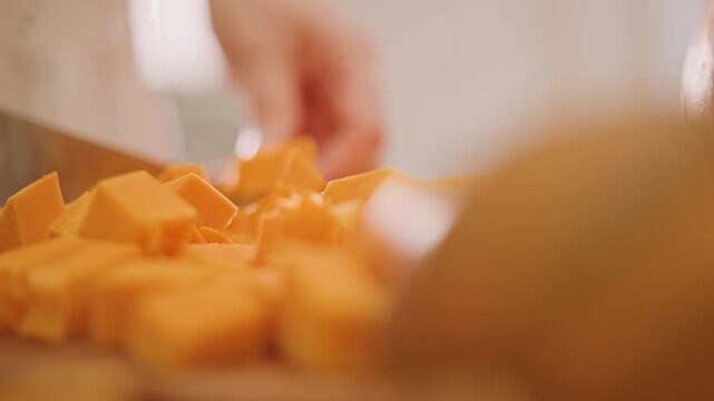 Closeup pumpkin cubes on cutting board, blade slicing edges with soft bokeh hand in background, golden pulp and seeds visible, artisan chef prepping precise pumpkin pie filling under warm studio