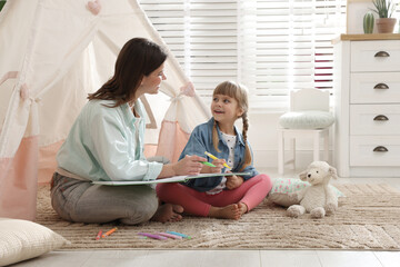 Mother and her daughter drawing in sketchbook together near toy wigwam at home, space for text © New Africa