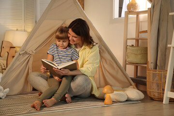 Mother and her daughter reading book near toy wigwam at home © New Africa
