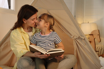 Mother and her daughter reading book near toy wigwam at home © New Africa