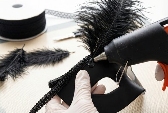 Gloved hands using a hot glue gun to attach black feathers to a decorative masquerade mask