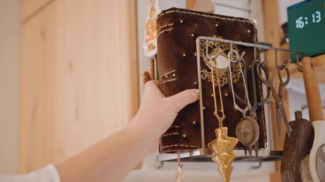 Velvet journal and heirloom jewelry, closeup of hand retrieving ornate book from metal rack by wooden cabinet, brass pendulum and layered chains drape across cover, warm ambient light, cozy rustic