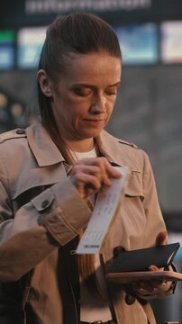 Woman Stands in Airport Terminal, Scanning Digital Ticket on Smartphone, Glances Between Phone Screen and Large Flight Information Display Board FIDS Confirm Gate and Departure Status. Vertical Shot.