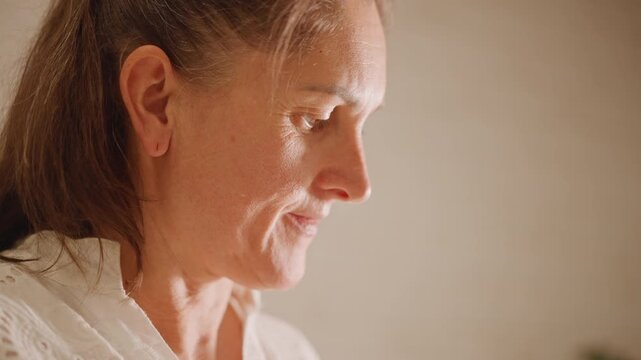 Closeup profile white woman reflecting soft side light subtle smile quiet reminiscence about pumpkin pie eyes cast downward skin texture and gray hair warm ambient glow intimate domestic portrait