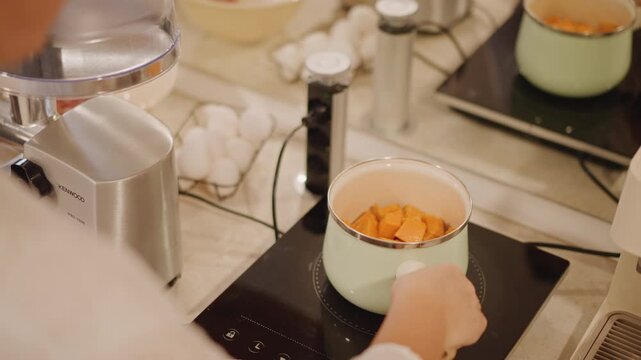 Pumpkin chunks simmering in saucepan on induction hob, blender and eggs visible, pastry chef adjusting heat, closeup on golden cubes breaking down, precise timing and modern stainless appliances,