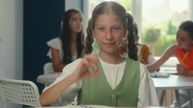 Positive cheerful Caucasian teenager kid child girl smiling demonstrated okay symbol pupil schoolgirl in school writing prepare exams write enjoying schoolwork education success showing ok in library