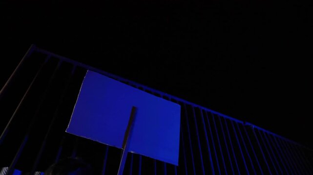Night protest footage. Back view of moving sign. Light flashes like sirens. Urban streets, cinematic tension. Anonymous protester, text hidden. Dark, moody, dramatic motion scene.