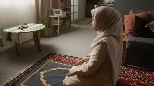 Cinematic zoom in shot of Muslim young woman wearing modest clothing and performing daily prayer ritual with sujud gesture on prayer mat, copy space