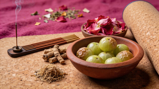 Wellness still life with fresh amla fruits in a clay bowl, ashwagandha roots, incense sticks, and rose petals on a cork yoga mat background