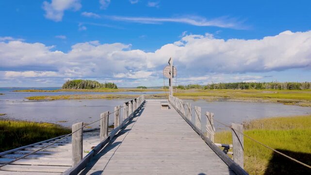 Slow motion footage of a wooden walkway across coastal marsh at Cape Forchu in Nova Scotia Canada leading towards a structure with water and distant land under blue sky with clouds in the summer time