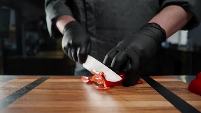 A chef in the kitchen is slicing a red pepper with a knife, his hands are wearing gloves.