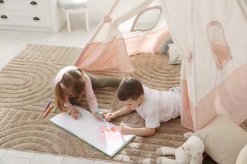 Little kids drawing in sketchbook together near toy wigwam at home, above view © New Africa