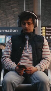 Man Sits in Airport Terminal, Wearing Large Silver Over Ear Headphones. Guy Listens Music, Scrolling Through Smartphone and Eventually Lifts Device Take Selfie or Record Video Message. Vertical Shot.