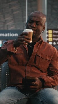 Man Sits in Airport Lounge, Using Smartphone Scan QR Code on Ticket. While Waiting Flight, Male Traveler Drinks Coffee, Calmly Double Checks Departure Details. Modern Travel Technology. Vertical Shot.