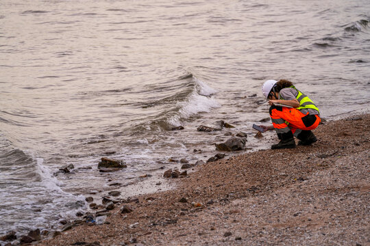 Female environmental scientist squats near shoreline, using smartphone and tablet to record water pollution data, highlighting environmental monitoring and coastal research.