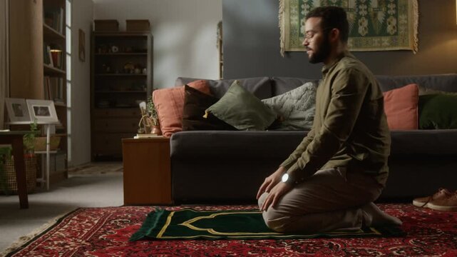 Cinematic side view shot of young Muslim man performing daily praying ritual on prayer mat in modern home with traditional elements, copy space