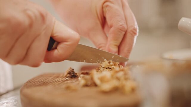 Closeup hands slicing walnuts on board, knife motion, warm kitchen light, preparing crunchy topping for pumpkin pie, shallow depth, cozy autumn baking scene, rustic texture and careful preparation