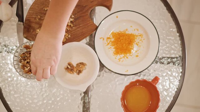 Topdown kitchen view with pumpkin zest, hands arranging walnuts and zest on plate, warm light on glass table, mug of juice nearby, intimate cozy atmosphere, methodical recipe staging