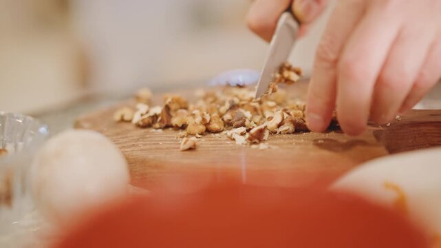 Hands chopping walnuts wooden board pumpkin pie garnish, knife skill closeup, red mixing bowl foreground, dough ball blurred, scattered crumbs and texture, rustic countertop and seasonal dessert