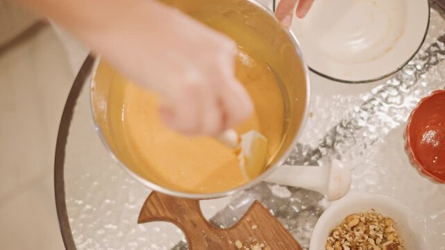 Mixing pumpkin pie filling in bowl, smooth orange batter stirred with spatula on glass table, bowls of nuts and ramekin visible, warm autumn lighting, cozy home kitchen, hands of baker preparing