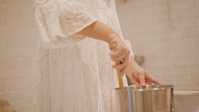 Hands stirring batter in bowl, wooden spoon folding puree and spices in stainless mixing bowl, side view of home baker concentrating, rhythmic motion and warm kitchen shelves, ambient soft light