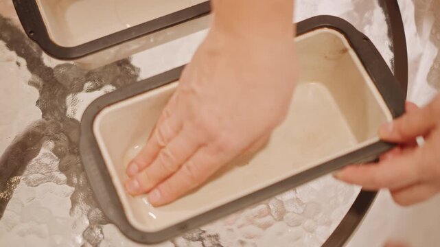 Hands greasing loaf pan for pumpkin loaf, palm rubbing oil across ceramic tin, two rectangular pans on textured glass table, even spread for nonstick finish, warm harvest baking atmosphere