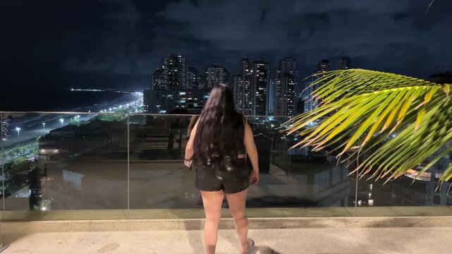 Modern lifestyle scene with woman watching oceanfront skyline at night in Barra Tijuca Brazil look down camera full body