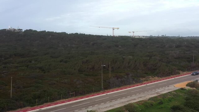 A calm drive along a scenic road winds through lush greenery. In the distance, cranes signify ongoing development, blending nature and progress beautifully.