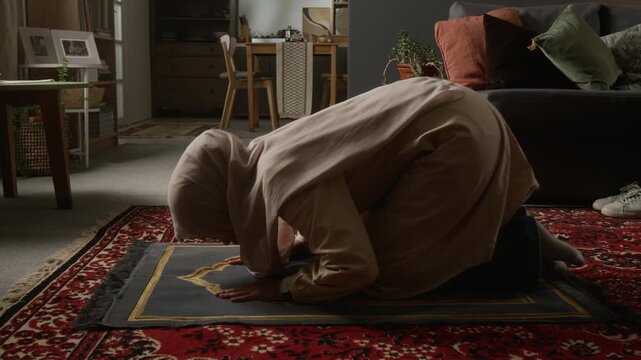 Side view shot of Muslim young woman performing sujud on prayer mat during daily praying ritual in modern home living room, copy space