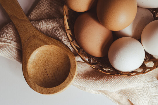 Farm fresh eggs in a rustic woven basket with a natural wooden spoon on a beige textile.Healthy nutrition and baking ingredients