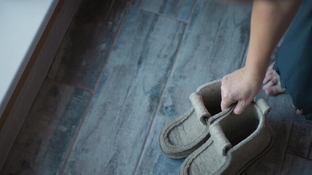 Cozy home morning, woman with slippers near window picks up pair, slips feet into soft wool slippers on wooden plank floor beside woven basket. Quiet routine, gentle light, relaxed posture, slow steps