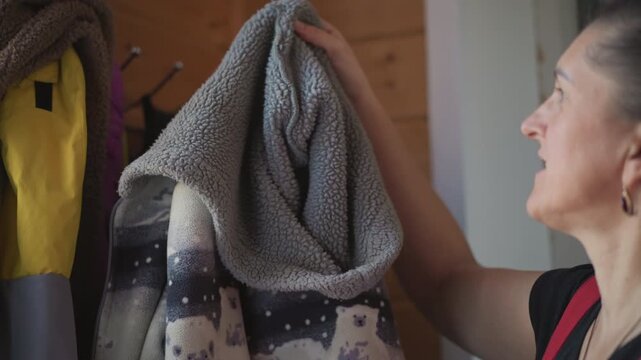 Woman at home putting on coat, adjusting fleece hood near wooden hallway hooks, gentle morning routine preparing to step outside, soft patterned fabric and practical outerwear, candid profile